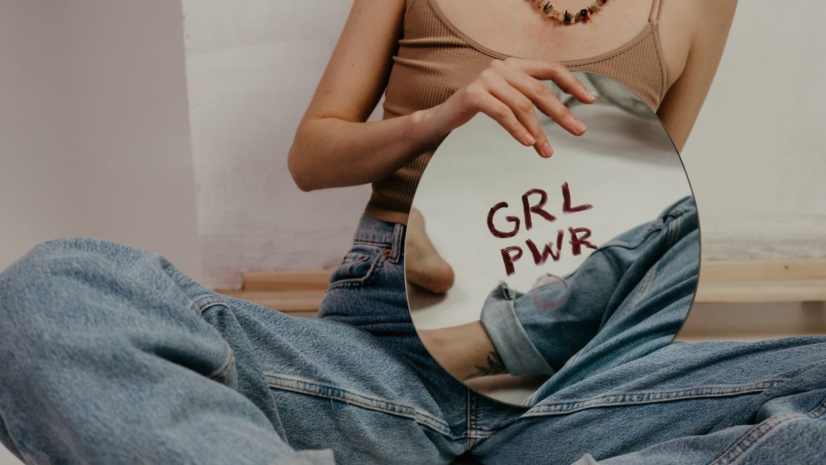 Young woman in jeans holding a mirror with 'GRL PWR' written, symbolizing empowerment.